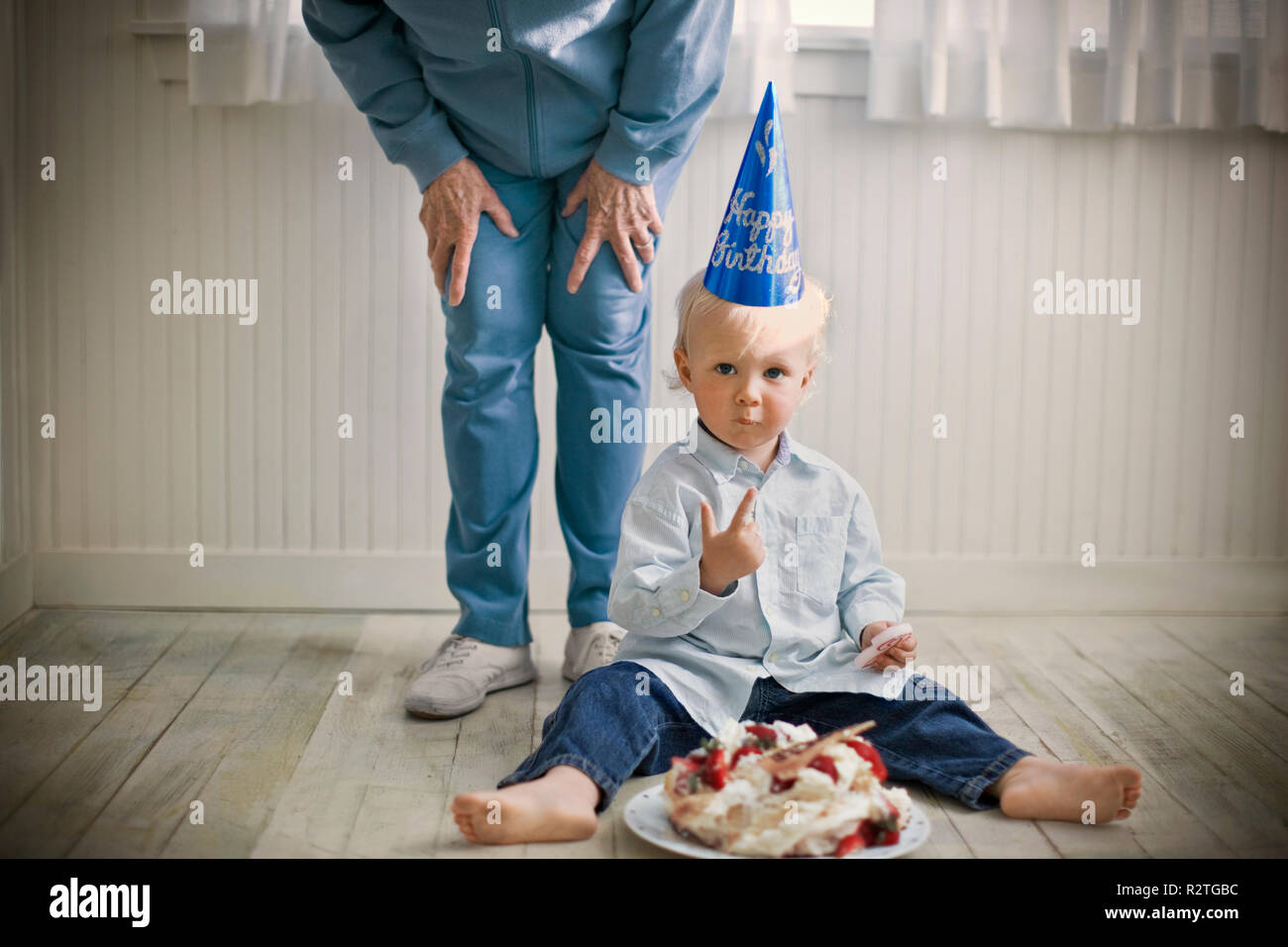 Ritratto di un giovane bambino seduto con i resti di una torta di compleanno davanti a lui su un pavimento di legno. Foto Stock