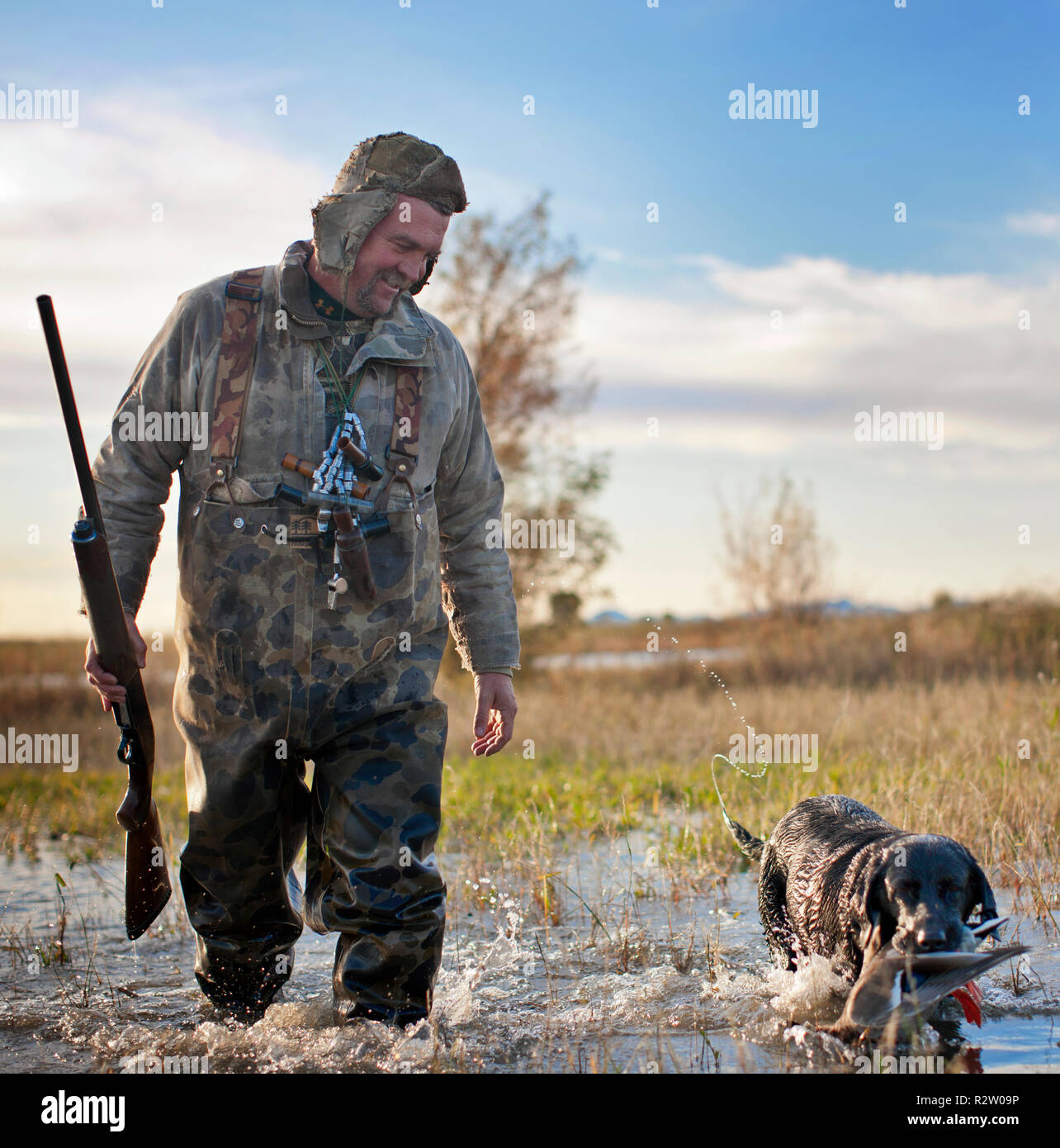 Cacciatore di anatre a piedi attraverso un lago poco profondo a fianco del suo cane che è in possesso di un anatra morta nella sua bocca. Foto Stock