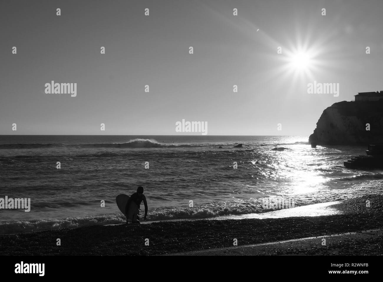Un surfista esce l'acqua a piedi fino alla spiaggia portando la sua tavola da surf a Freshwater Bay Isle of Wight. Un brillante sole risplende sulla scena wintery Foto Stock