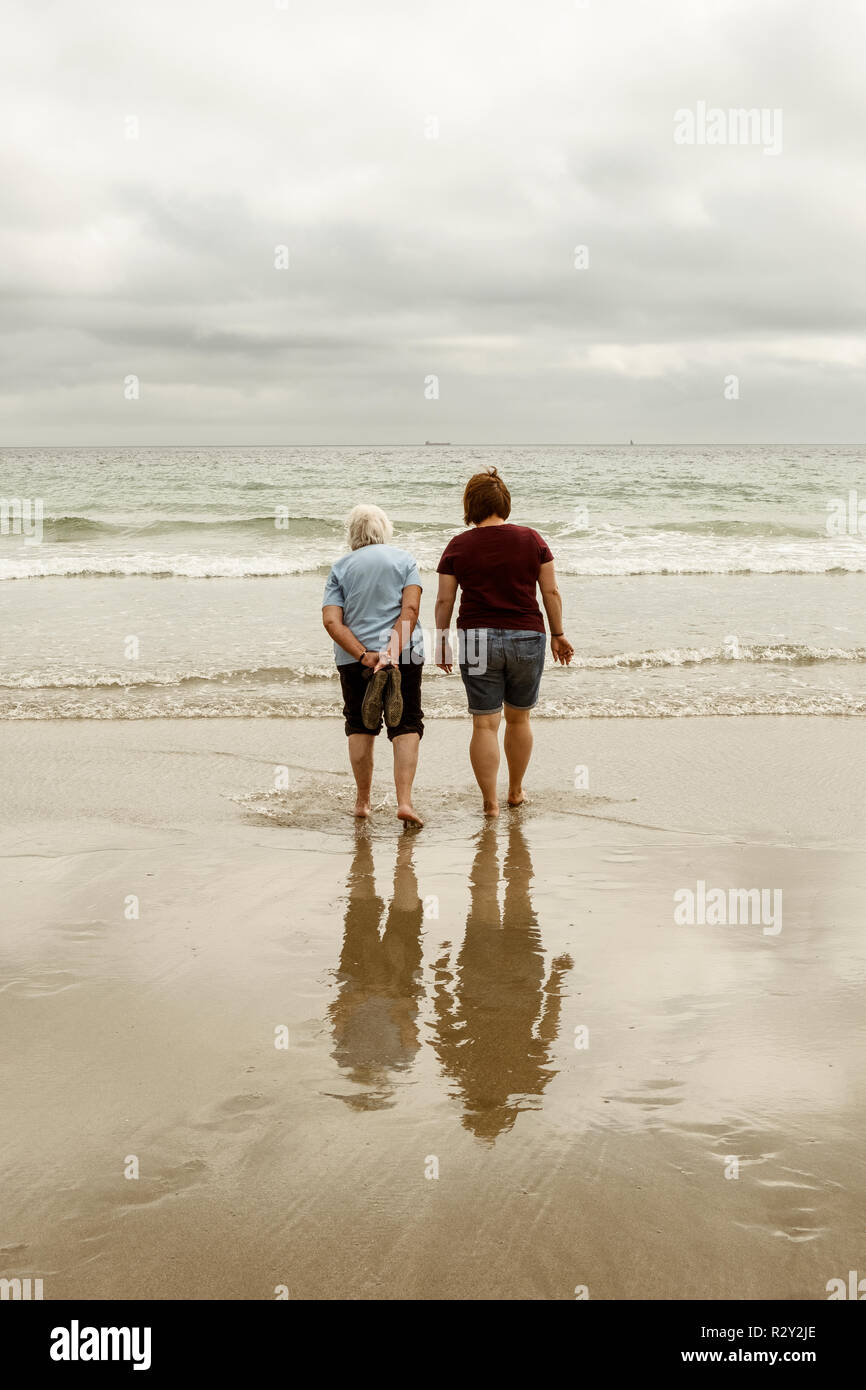 Vista posteriore di un pelo grigio donna anziana e una giovane donna sguazzare con le scarpe fuori in ondulazioni su di una spiaggia di sabbia. Foto Stock