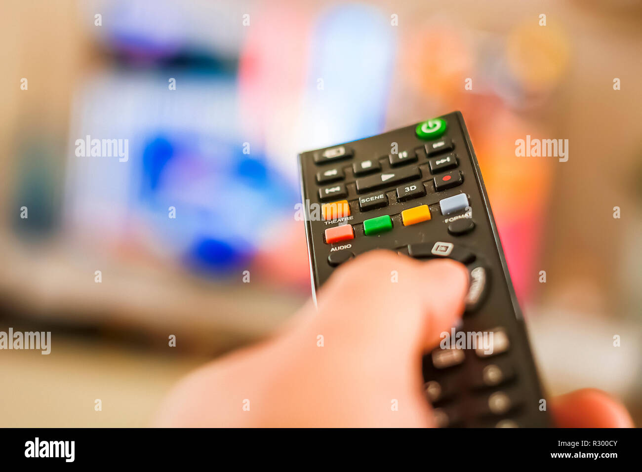 Una mano premendo un tasto del telecomando per cambiare i canali di fronte a una sfocata sullo schermo della televisione Foto Stock