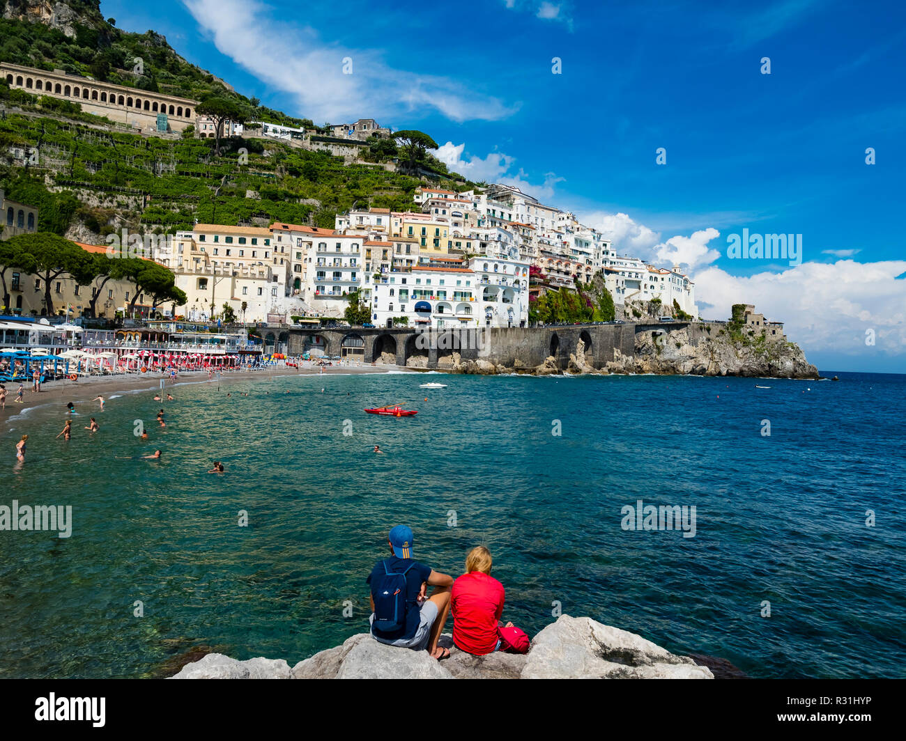 Giovane si siede sulla roccia e guarda la città vecchia e la Spiaggia di Amalfi, penisola di Sorrento, Costiera Amalfitana, Campania, Italia Foto Stock