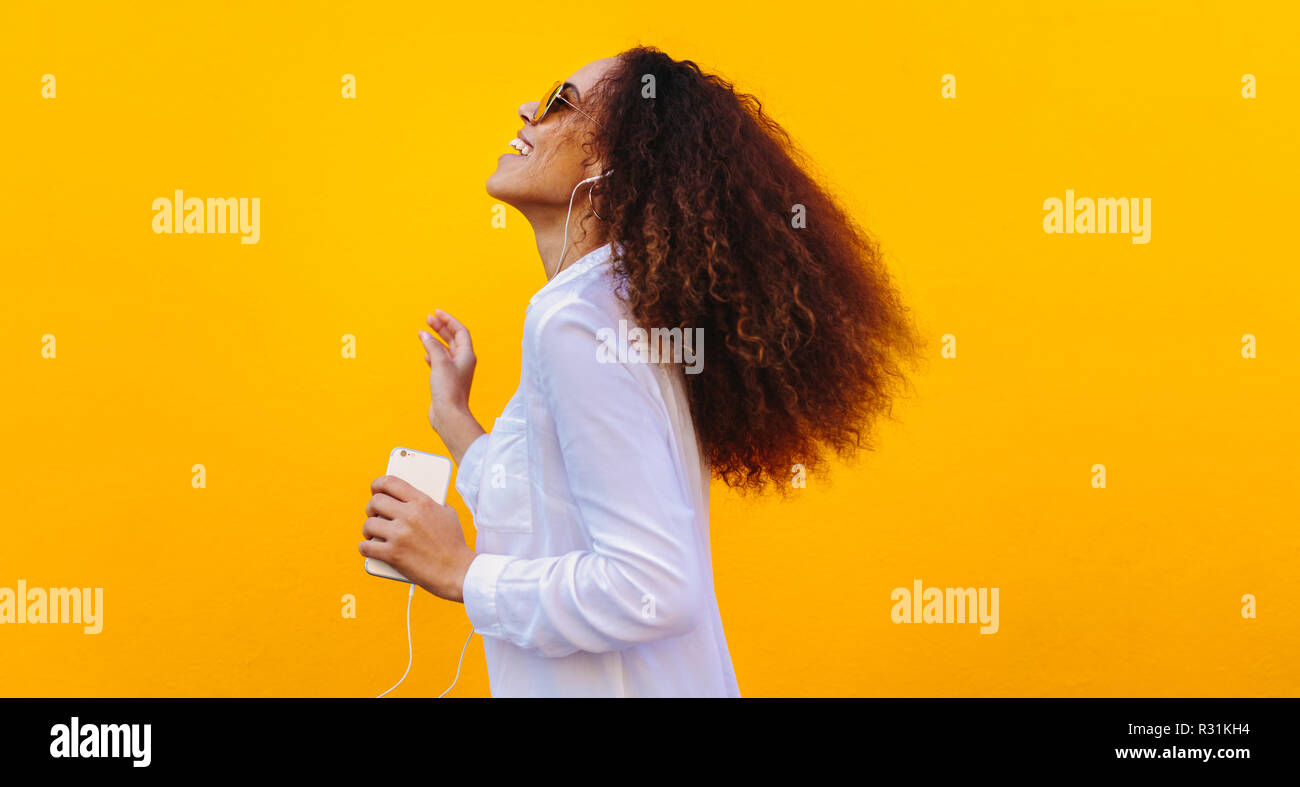 Vista laterale della donna che indossa gli auricolari per ascoltare la musica dal suo telefono cellulare. Ragazza africana avendo divertimento mentre si ascolta la musica sullo smartphone. Foto Stock