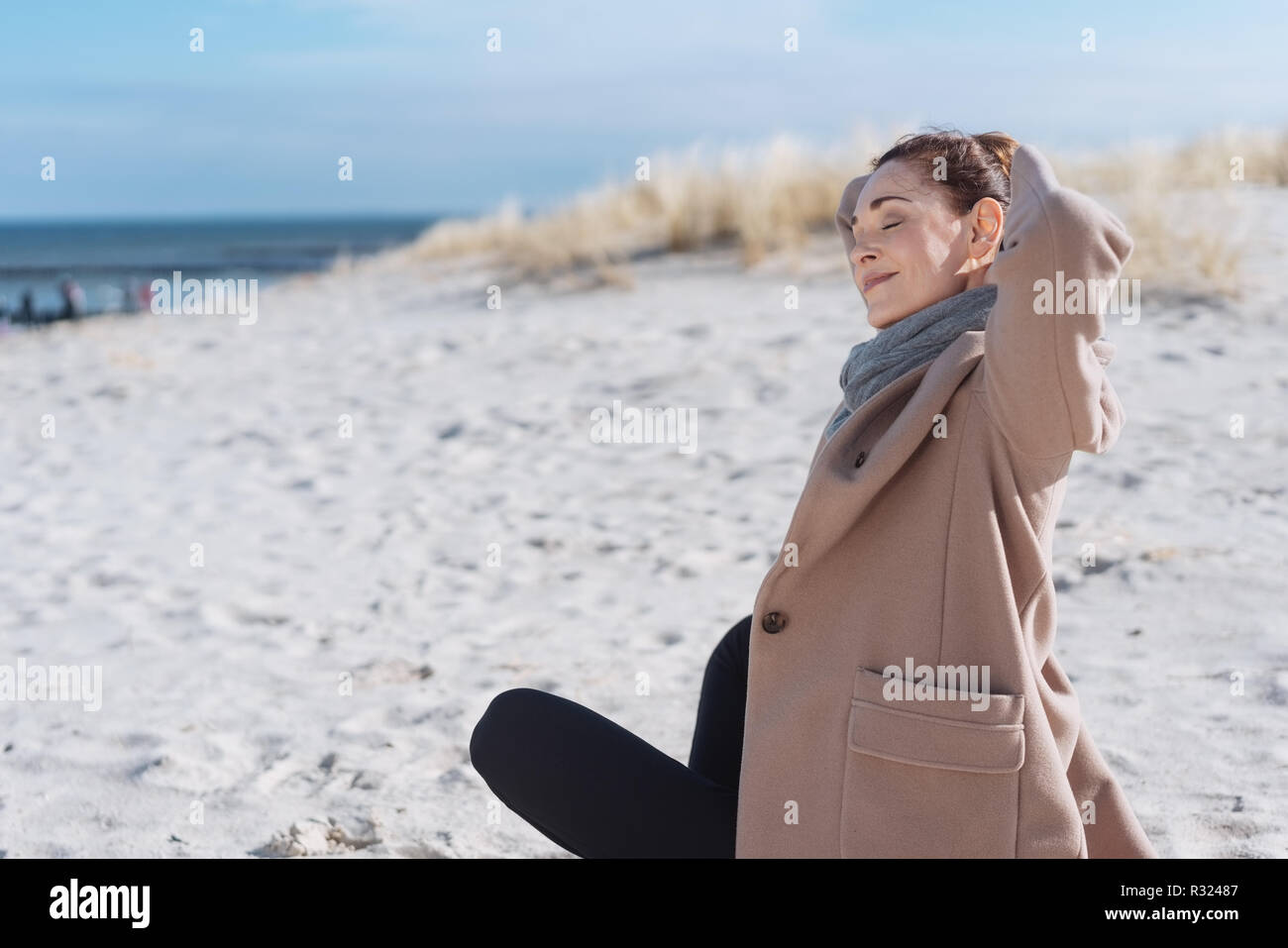 Giovane donna trascorrere una rilassante giornata invernale in una spiaggia a suo spesso mantello e sciarpa seduti sulla sabbia stretching le braccia con un sorriso di felicità Foto Stock