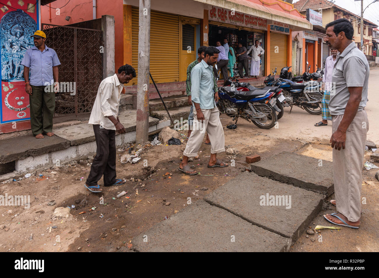 Belur, Karnataka, India - 2 Novembre 2013: cinque ragazzi di cui calcestruzzo pesante piastra per coprire una linea di cucitore. Scena di strada con la gente e i motocicli. Foto Stock