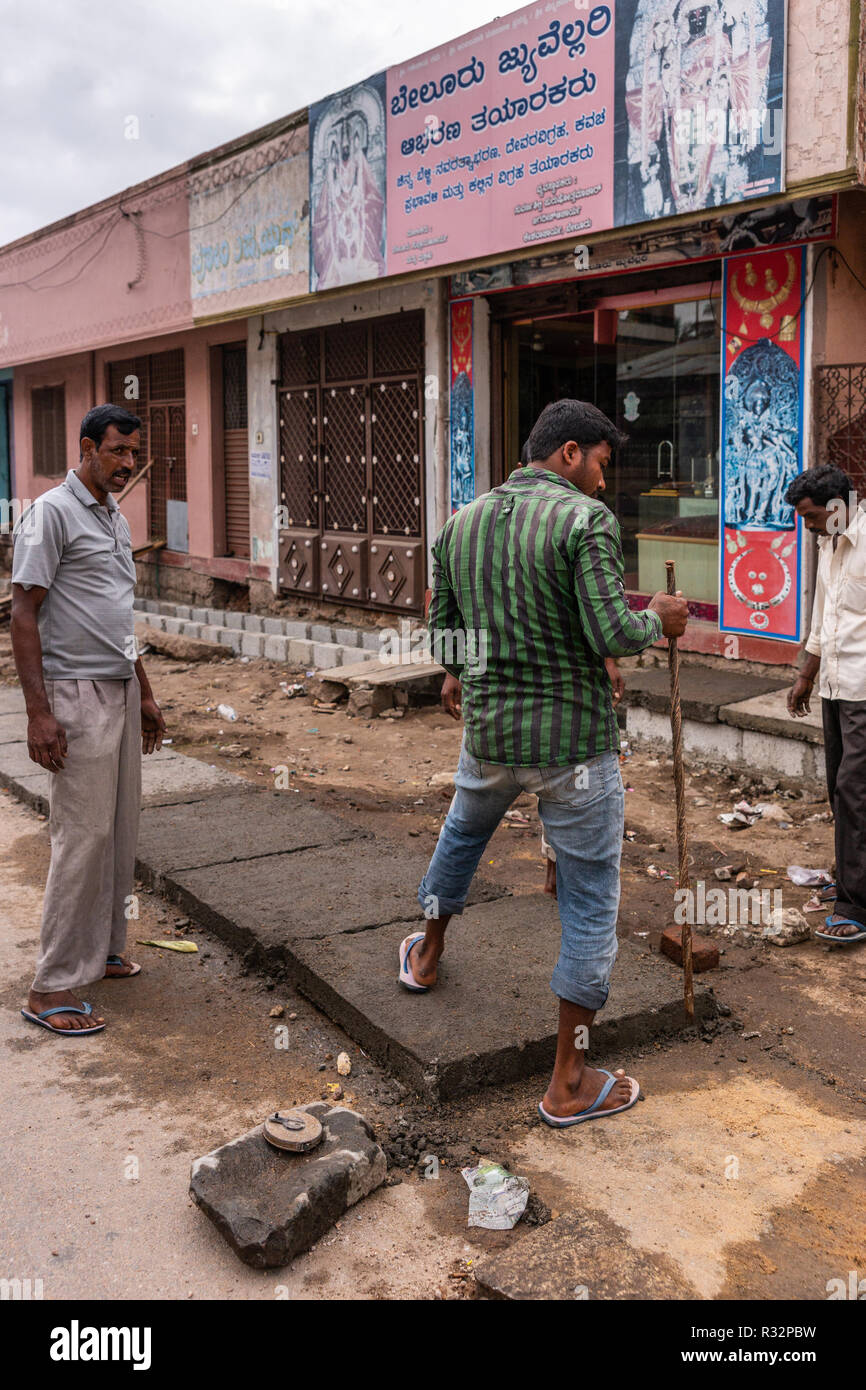 Belur, Karnataka, India - 2 Novembre 2013: tre ragazzi di cui calcestruzzo pesante piastra per coprire una linea di cucitore. Scena di strada con negozi e poster. Foto Stock