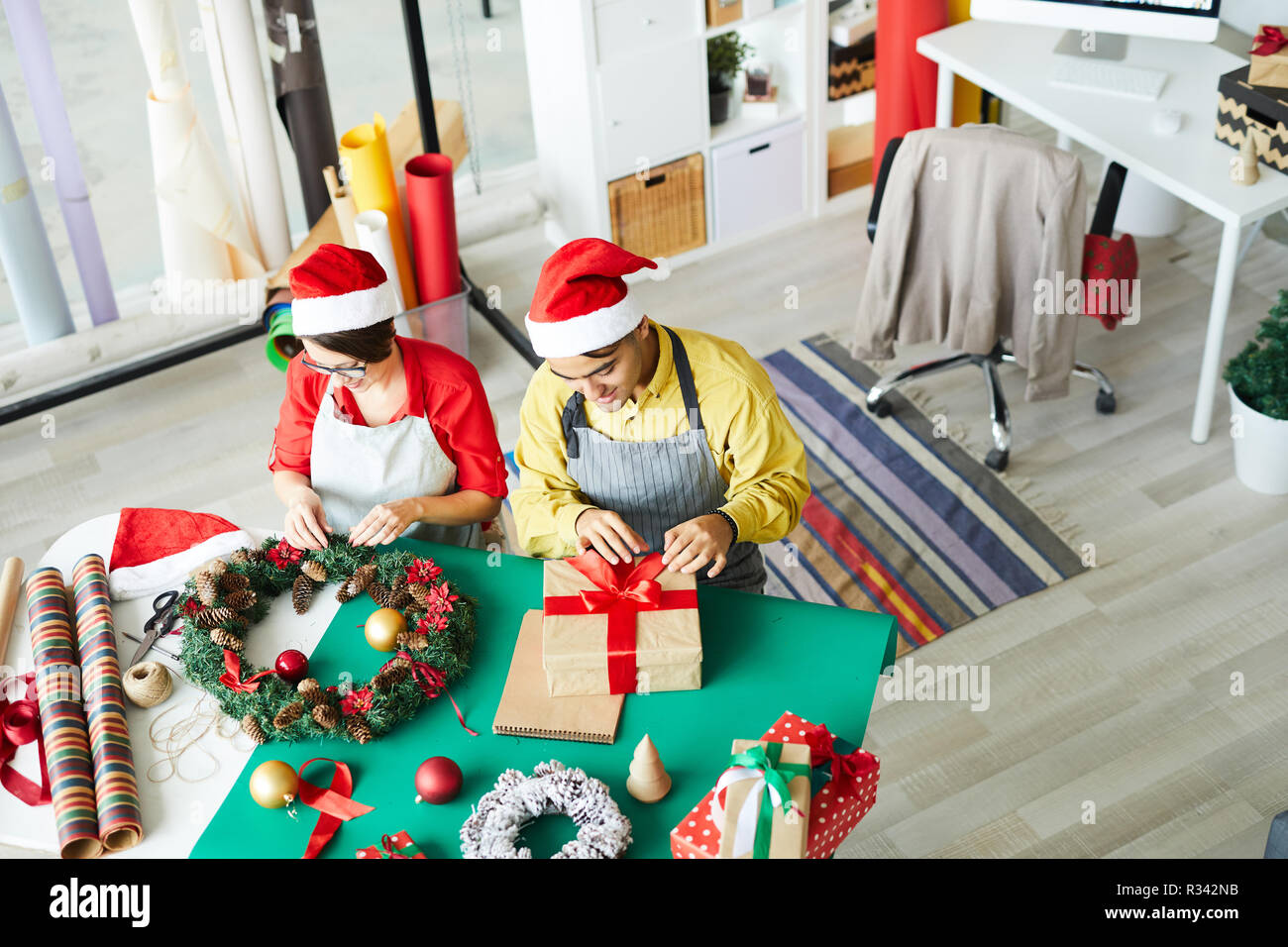 Due giovani colleghi in grembiuli e Santa caps lavorare sulle decorazioni di Natale e regali di imballaggio Foto Stock