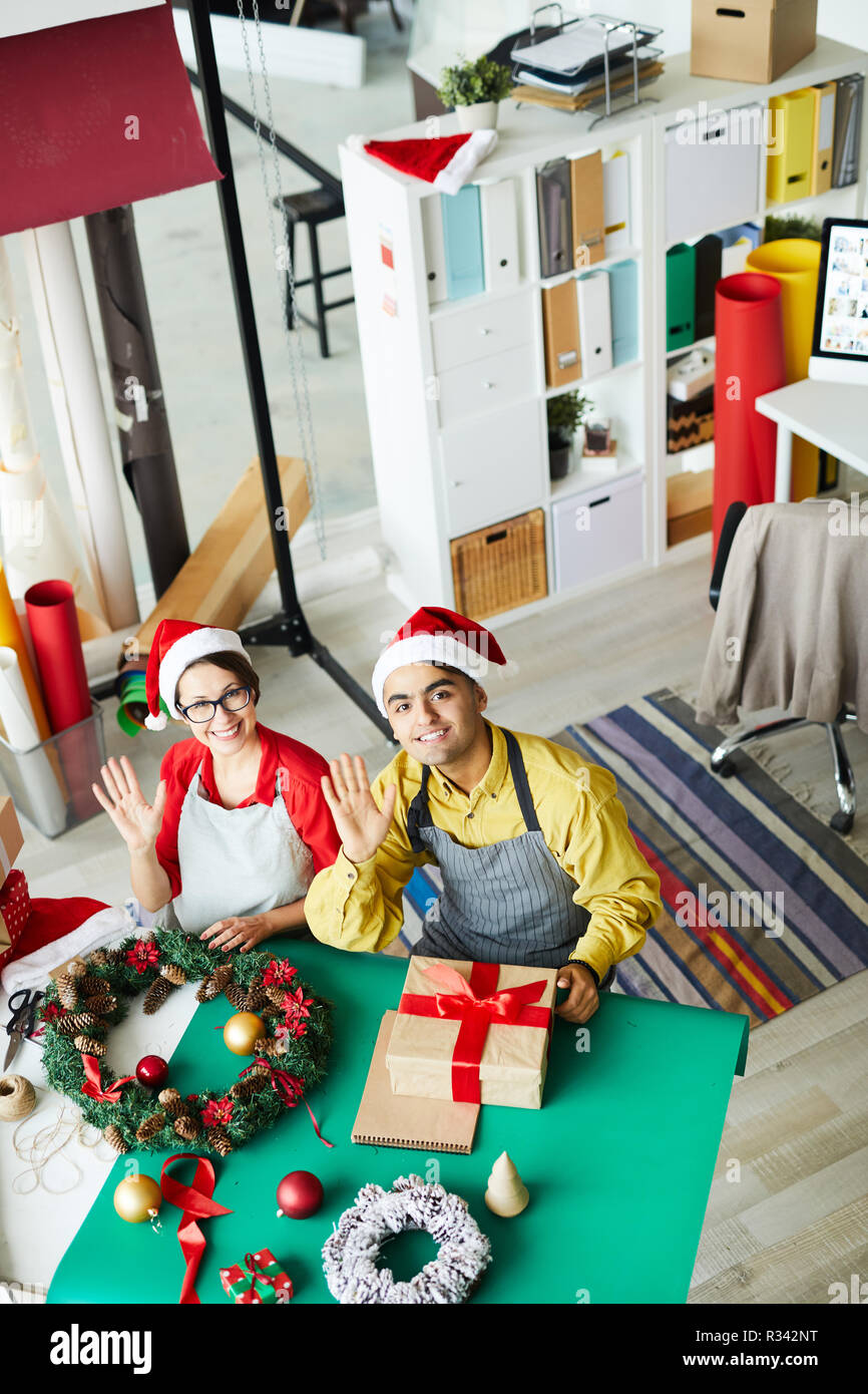 Felice l'uomo e la donna in abbigliamento da lavoro e Santa caps sventolare le mani mentre si preparano i regali di natale Foto Stock