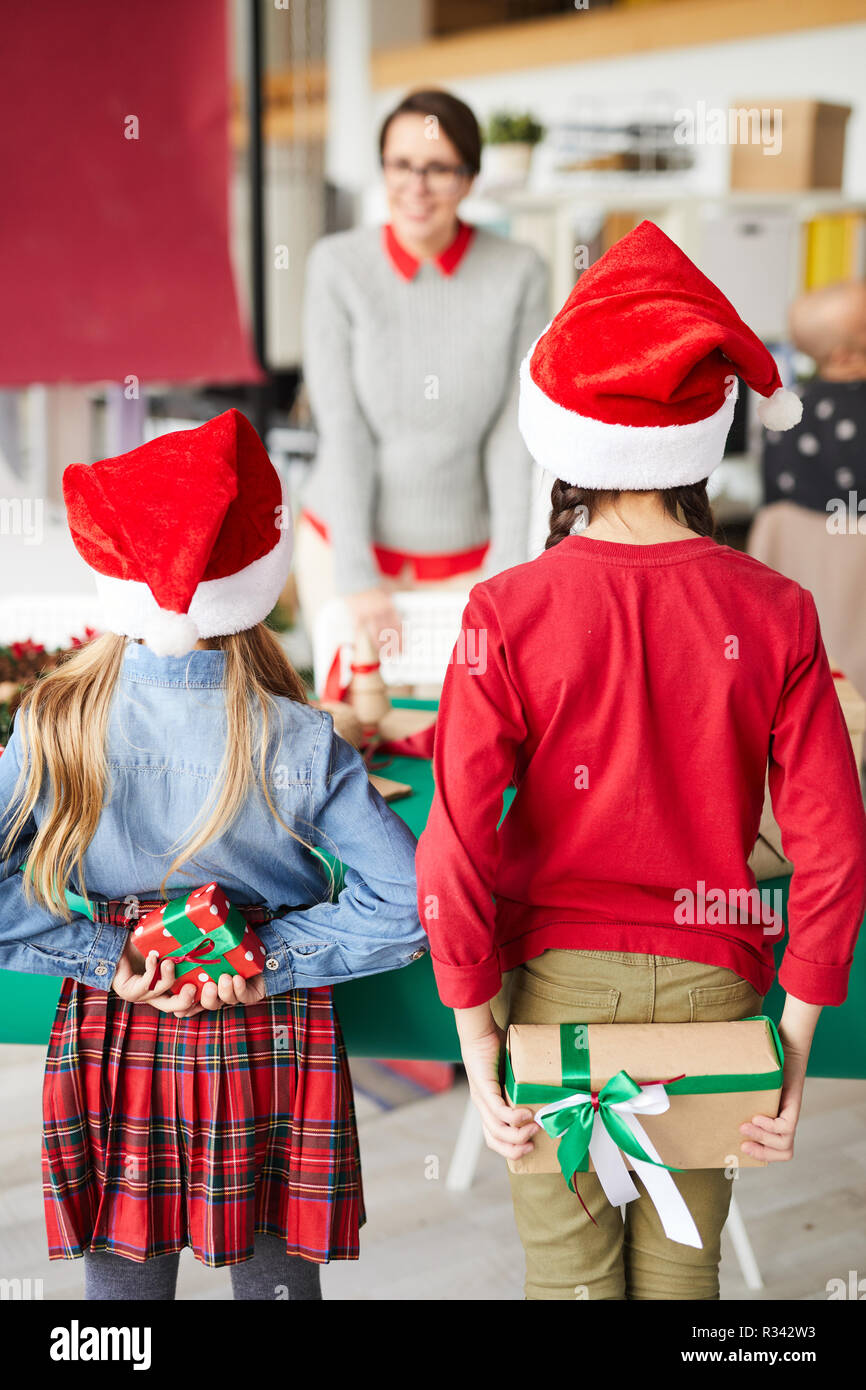 Vista posteriore di due bambine in Santa caps nascondere i regali di Natale per la loro madre dietro spalle Foto Stock