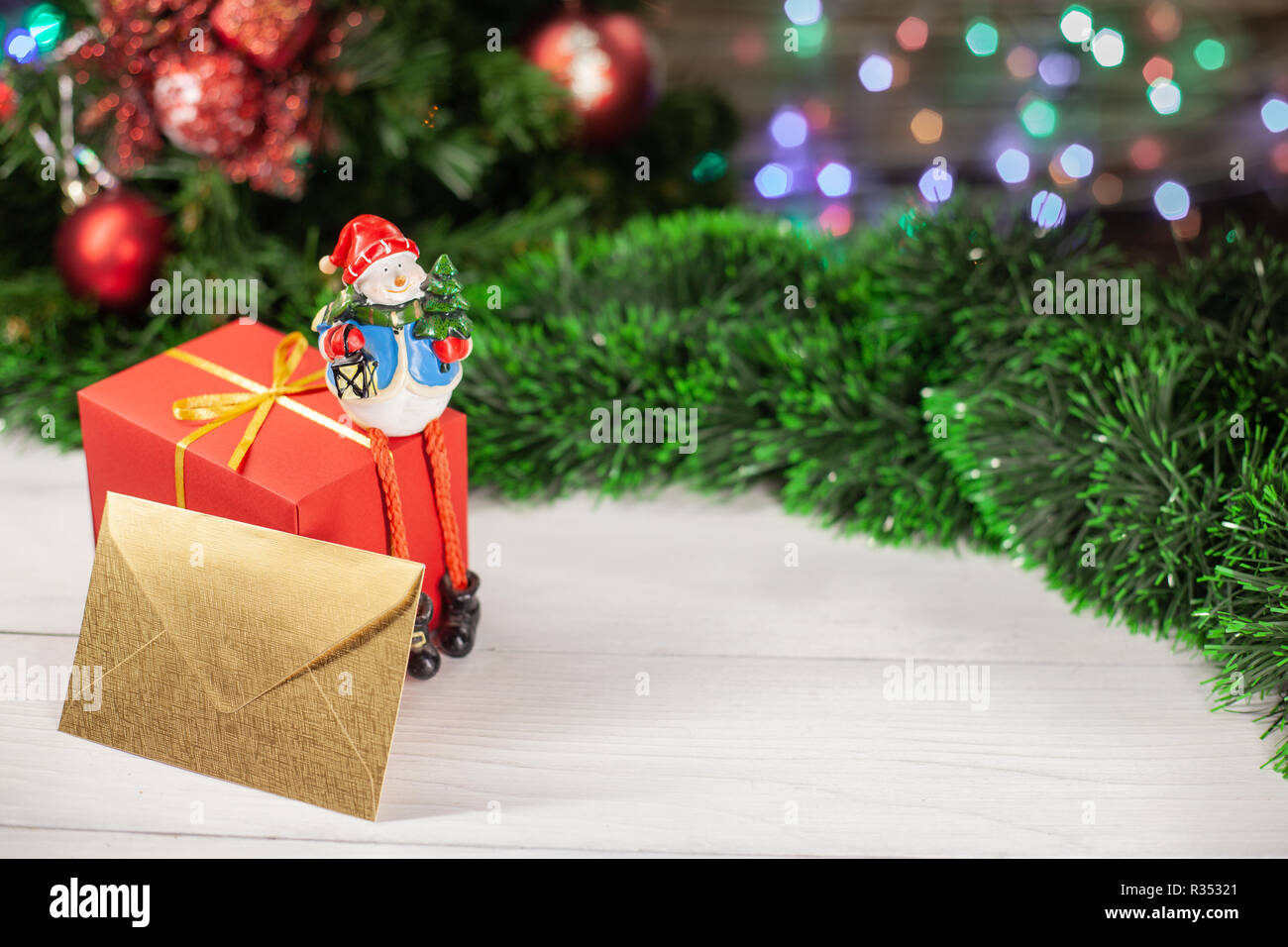 Natale la figura di un pupazzo di neve seduti su un regalo in carta rossa accanto a una busta di oro che è su un bianco tavolo di legno accanto a un verde tinsel in ba Foto Stock