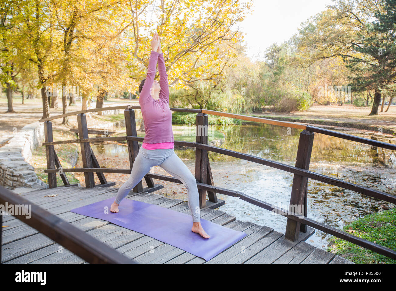 Giovane donna di eseguire esercizi Yoga nella posizione del Guerriero pongono sul ponte di legno al parco. Mattina giornata d'autunno. Una sana motivazione. Foto Stock