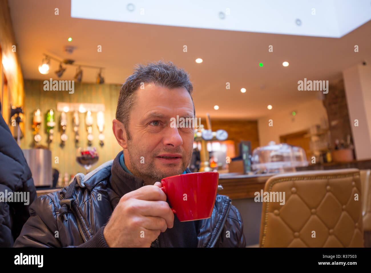 Primo piano di attraente maschio caucasico, nei suoi anni '40, all'interno di bar trendy godendo di un caffè da una tazza rossa che sta tenendo. Foto Stock
