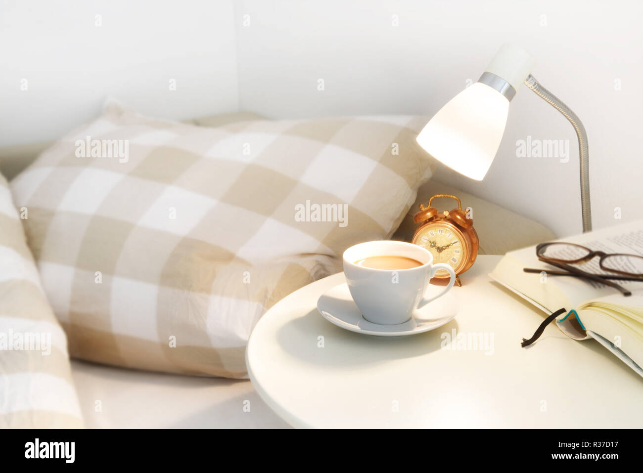 Il caffè al mattino sul comodino, più una sveglia, lampada e prenota con occhiali, giorno pigro concetto, selezionato soft focus, profondità di campo ridotta Foto Stock