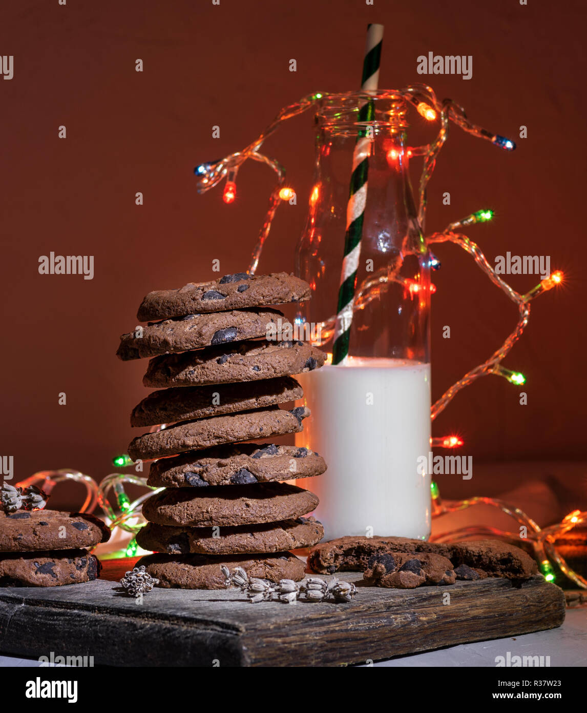 Pila di cioccolato biscotti rotondo, di una bottiglia di vetro con il latte e un luminoso ghirlanda di Natale, sfondo festivo Foto Stock