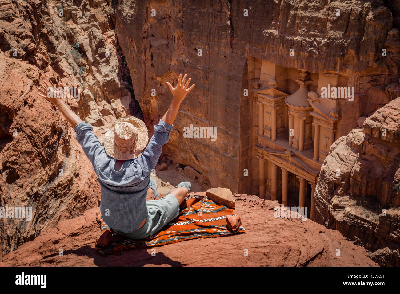 Turistico con cappello per il sole si siede con i bracci sollevati sul tappeto e guarda dall'alto nel canyon Siq, faraone treasure house scolpito Foto Stock