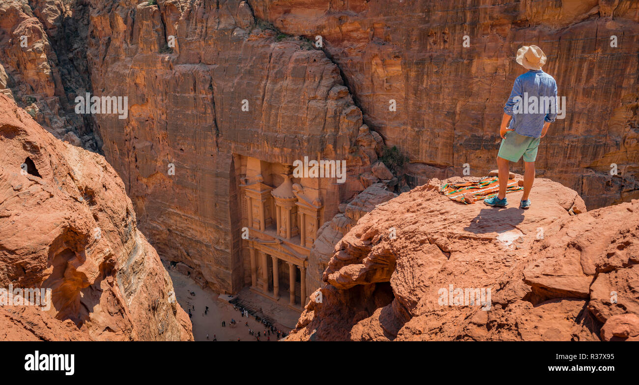 Turistico con cappello per il sole sorge su rocce e guarda dall'alto nella gola Siq, faraone treasure house scavata nella roccia, facciata Foto Stock