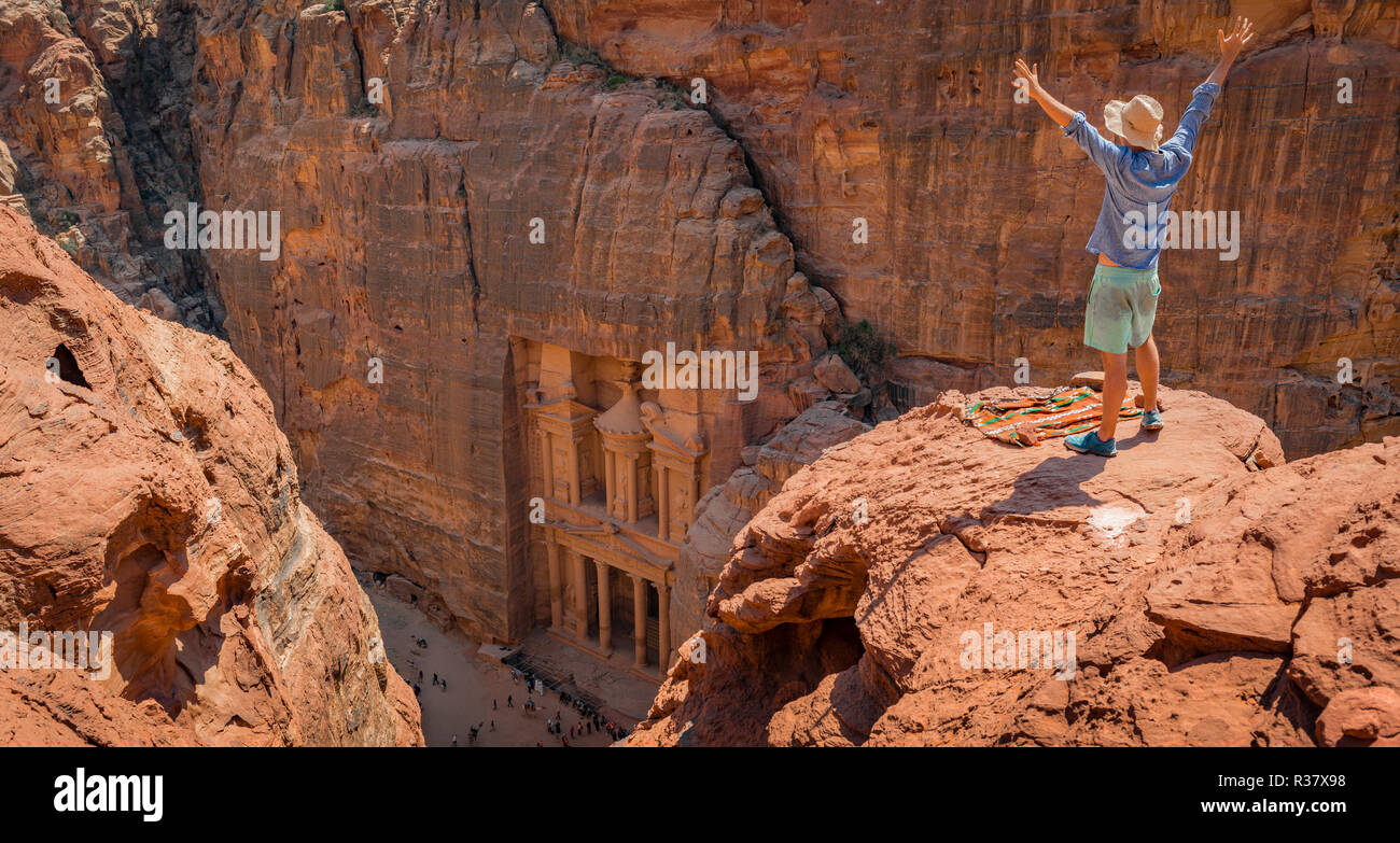 Turistico con cappello per il sole sorge su rocce con bracci sollevati e si guarda dal di sopra nella gola Siq, faraone treasure house scolpito Foto Stock