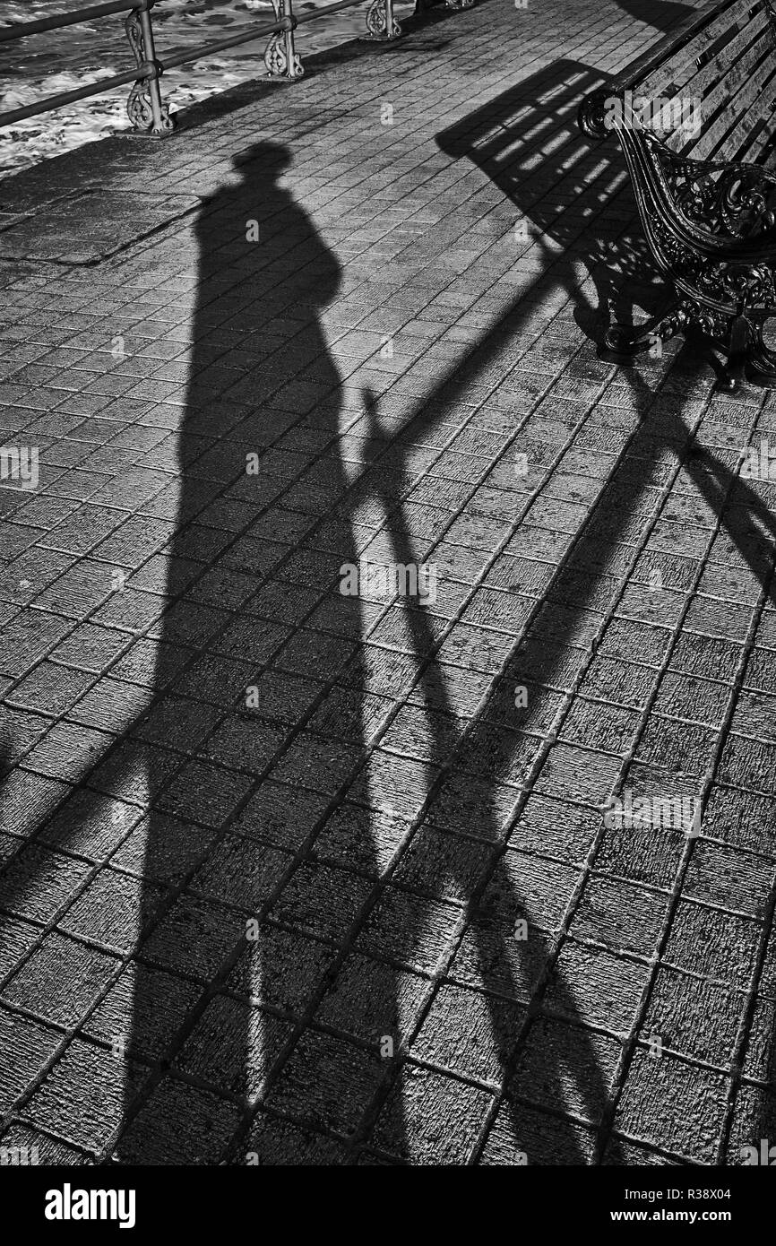 L ombra di un uomo che indossa un cappello sul marciapiede di un pontile realizzato da blocchi quadrati in bianco e nero Foto Stock