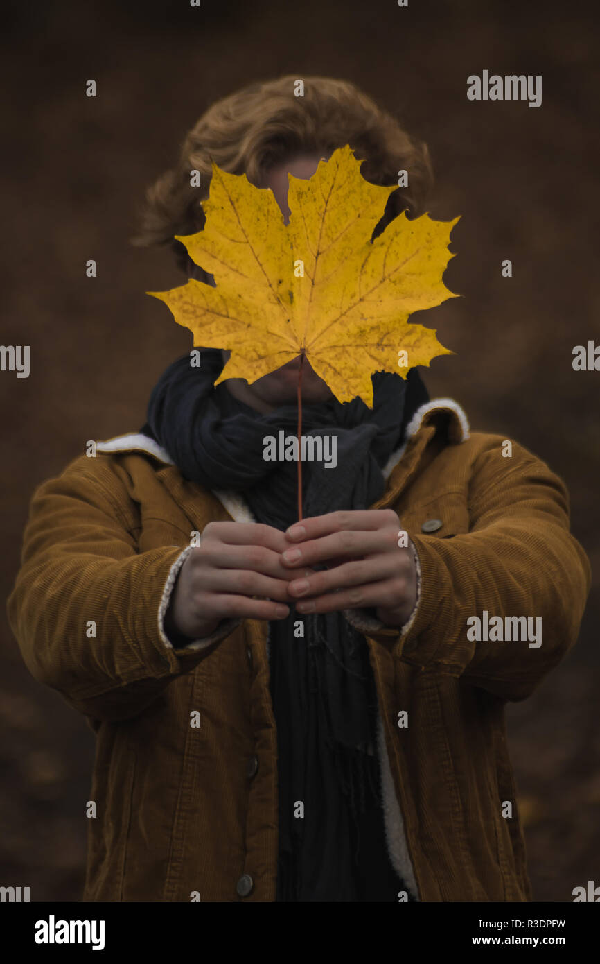 Un ritratto concettuale di un giovane maschio che tiene una foglia di acero gialla davanti al suo volto durante l'autunno in giacca coordinata Foto Stock