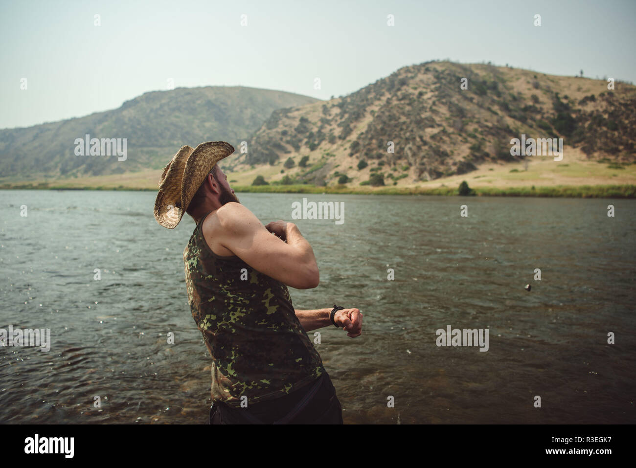 Candid fotografia di un uomo in un cappello di cowboy, saltando pietre attraverso il fiume come egli prende una pausa dalla guida sul suo viaggio per allungare le gambe. Foto Stock