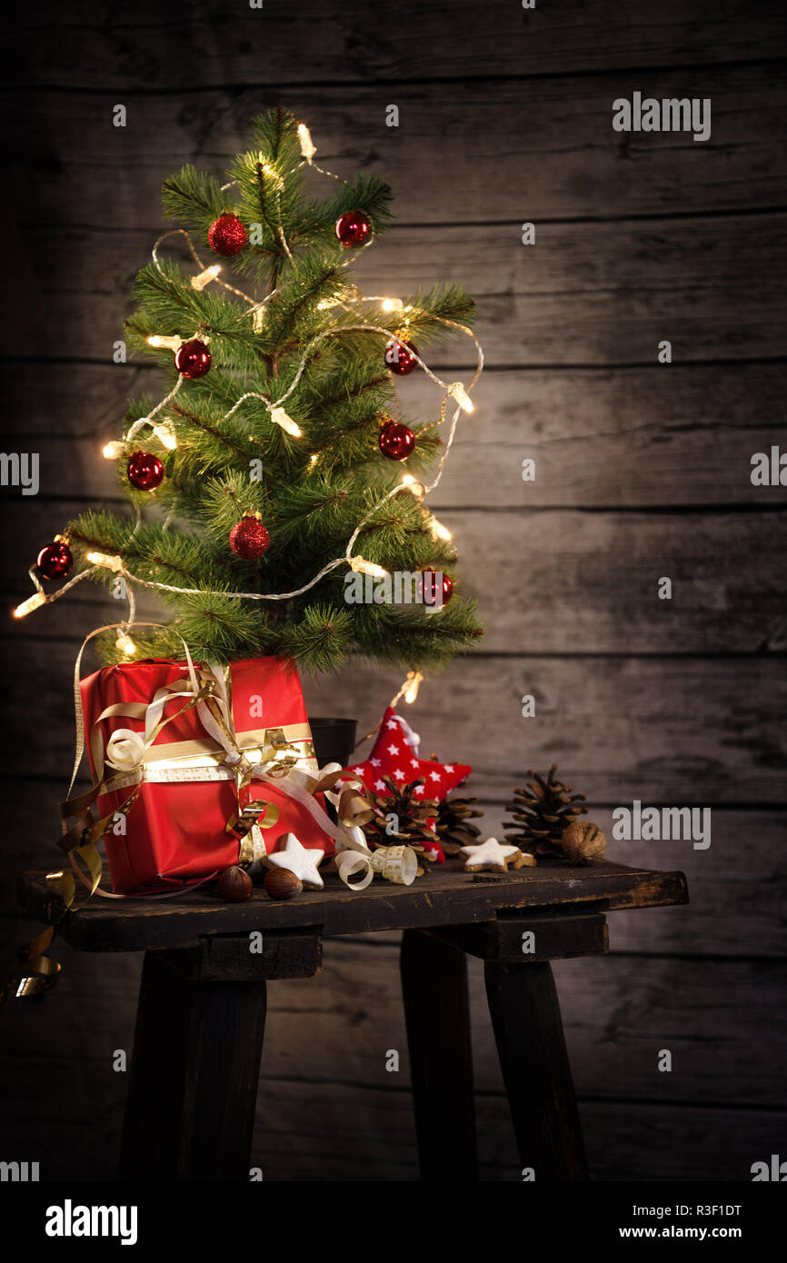 Piccolo albero di Natale artificiale con catena leggera, dono e decorazione su un vecchio sgabello davanti un rustico di pareti in legno, copia spazio, selezionato focu morbido Foto Stock