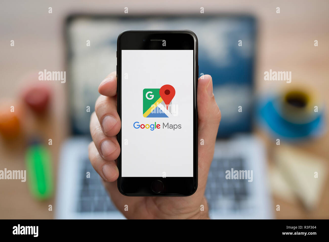 Un uomo guarda al suo iPhone che consente di visualizzare le mappe di Google logo, mentre se ne sta seduto alla sua scrivania per computer (solo uso editoriale). Foto Stock