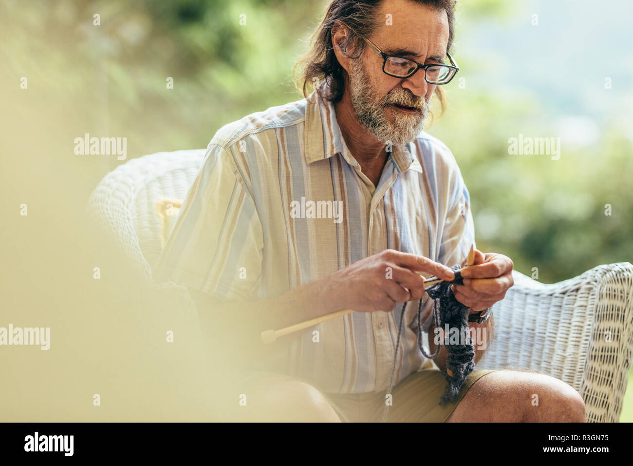 Uomo anziano con la barba seduto su una sedia e la maglia. Il vecchio uomo maglia con aghi e filati di lana. Foto Stock