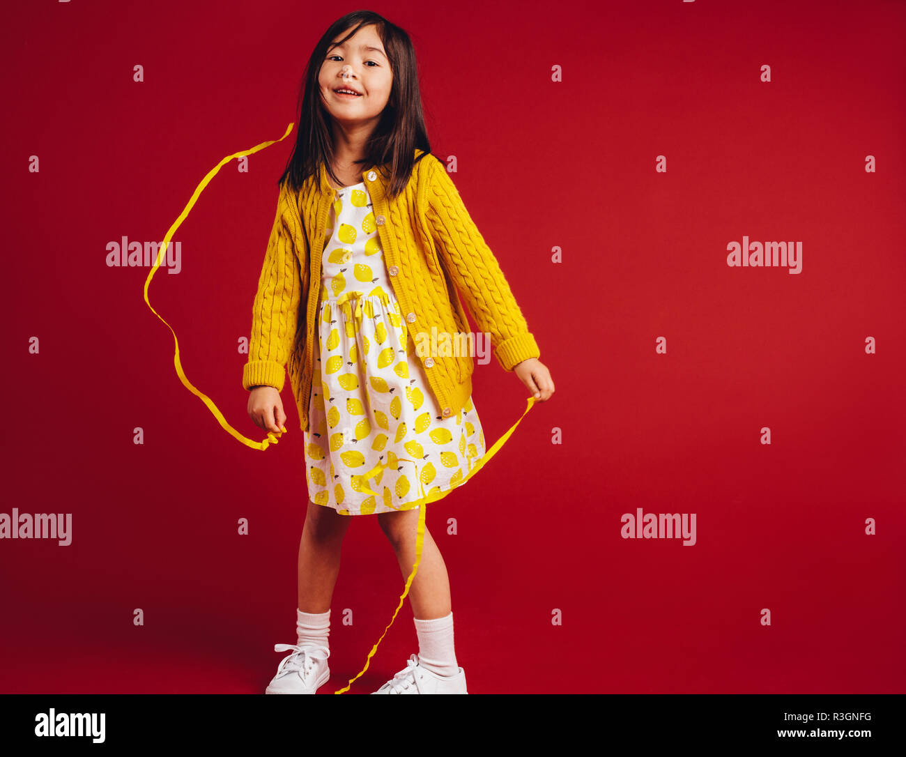 Sorridente asian kid sventolando nastri. Bambina giocando con giallo nastri di raso in piedi contro uno sfondo di colore rosso. Foto Stock
