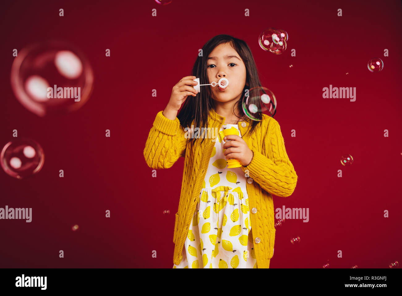 Asian kid a giocare con le bolle di sapone. Bambina in piedi contro uno sfondo rosso a soffiare bolle. Foto Stock