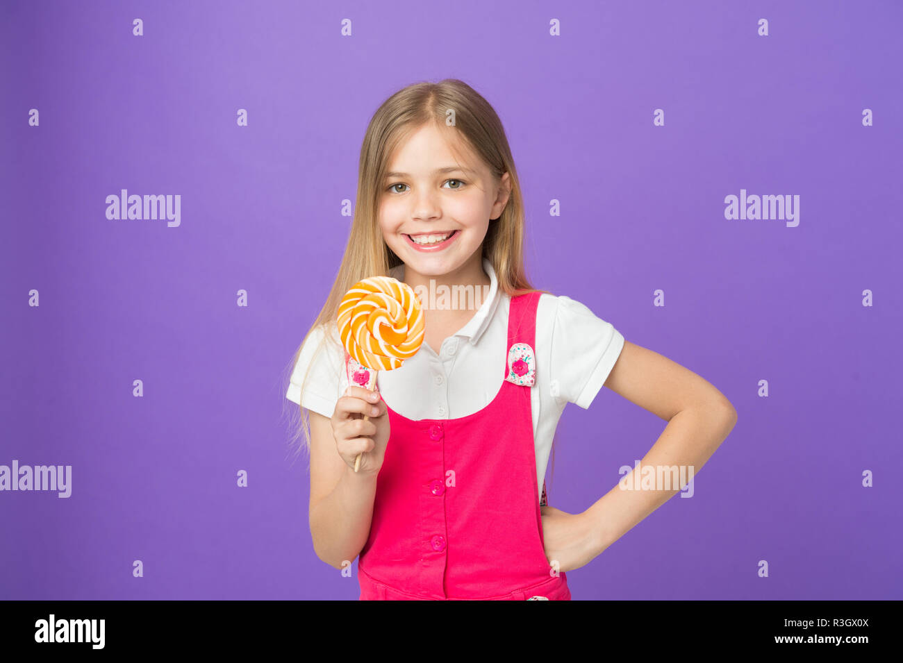 Bambino sorridente con luminosi lollipop. Ragazza con lunghi capelli biondi in abito rosa isolato su sfondo viola. Dente dolce con la faccia felice azienda big ro Foto Stock