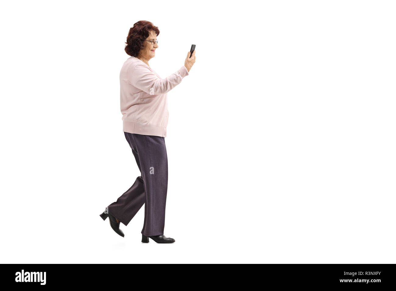 Lunghezza piena ripresa del profilo di una signora anziana a piedi con un telefono cellulare isolato su sfondo bianco Foto Stock