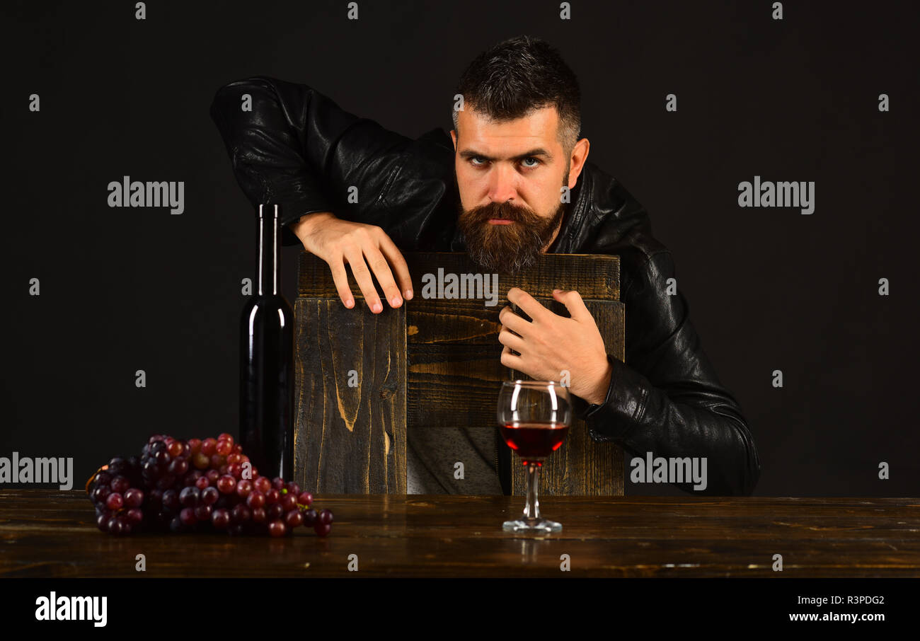 Sommelier si appoggia sulla sedia in legno. Uomo con la barba vicino a un bicchiere di vino sul marrone scuro dello sfondo. Degustator con gravi faccia per bottiglia per vino e grap scuro Foto Stock