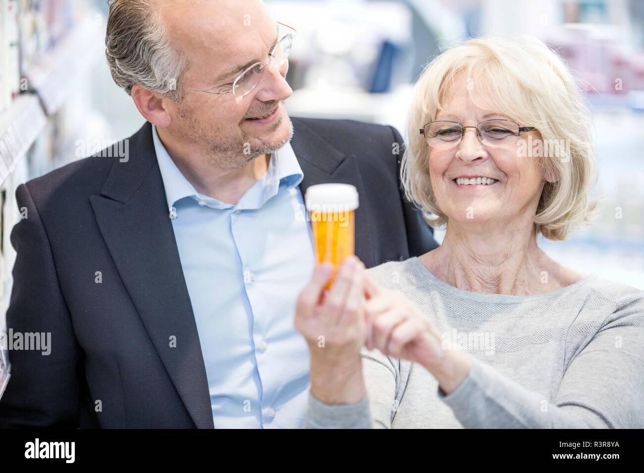 Sorridente donna senior che mostra una pillola etichetta del flacone di uomo maturo in farmacia. Foto Stock