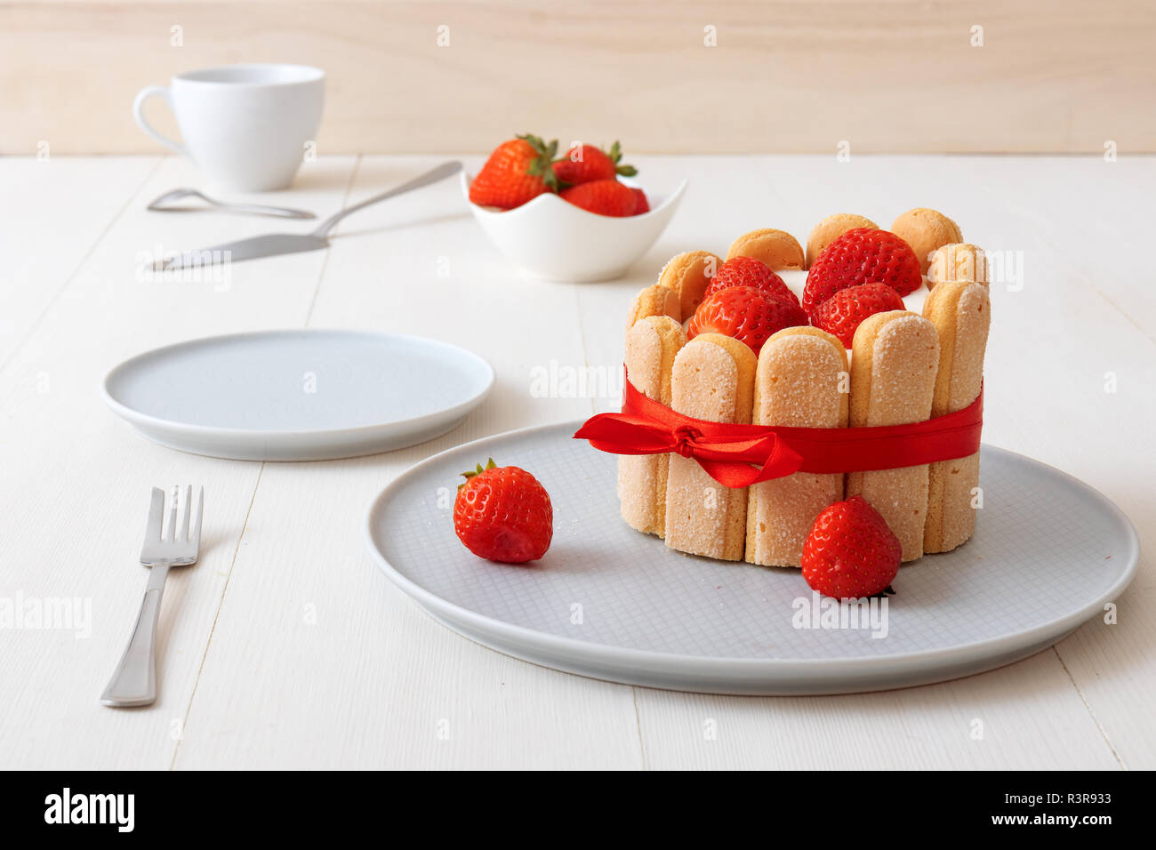 Charlotte, francese dessert con fragole, legato con un nastro rosso scarlatto, tazza di caffè, la ciotola con le fragole sul tavolo bianco. Foto Stock