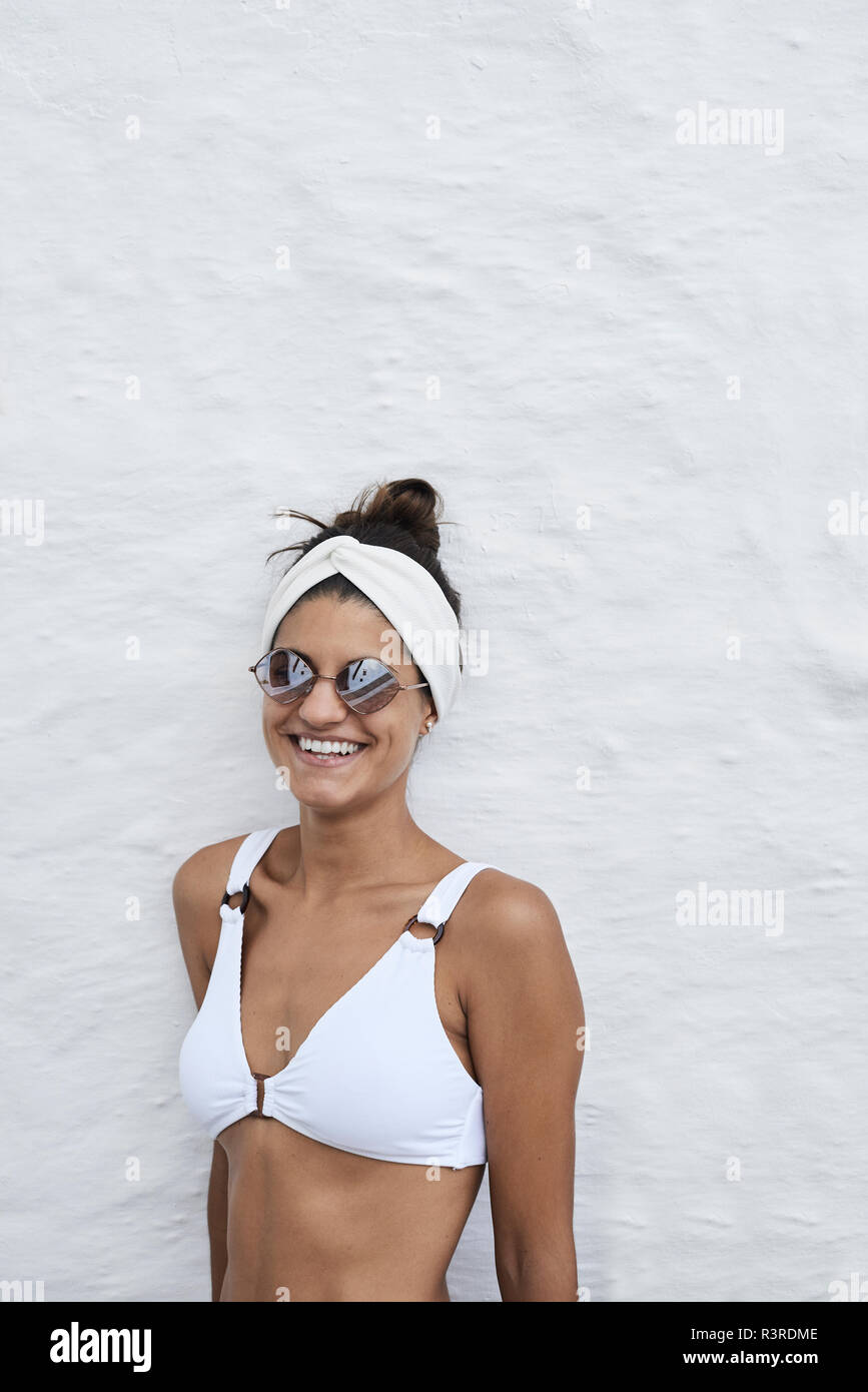 Ritratto di ridere giovane donna indossa bikini bianco top appoggiata contro il muro bianco Foto Stock