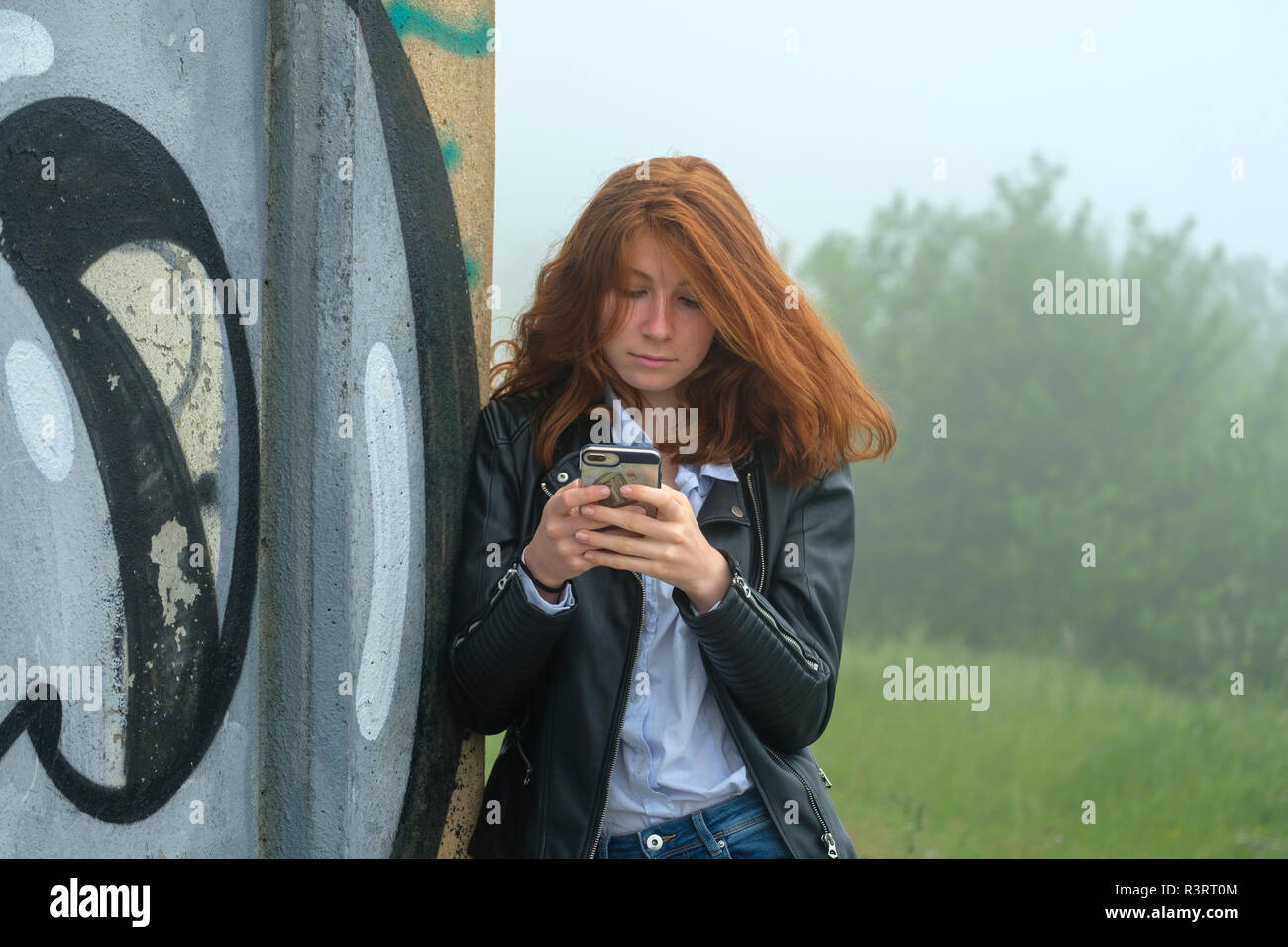 L'Italia, Finale Ligure, ritratto di testa rossa ragazza adolescente appoggiata contro il murale guardando al telefono cellulare Foto Stock