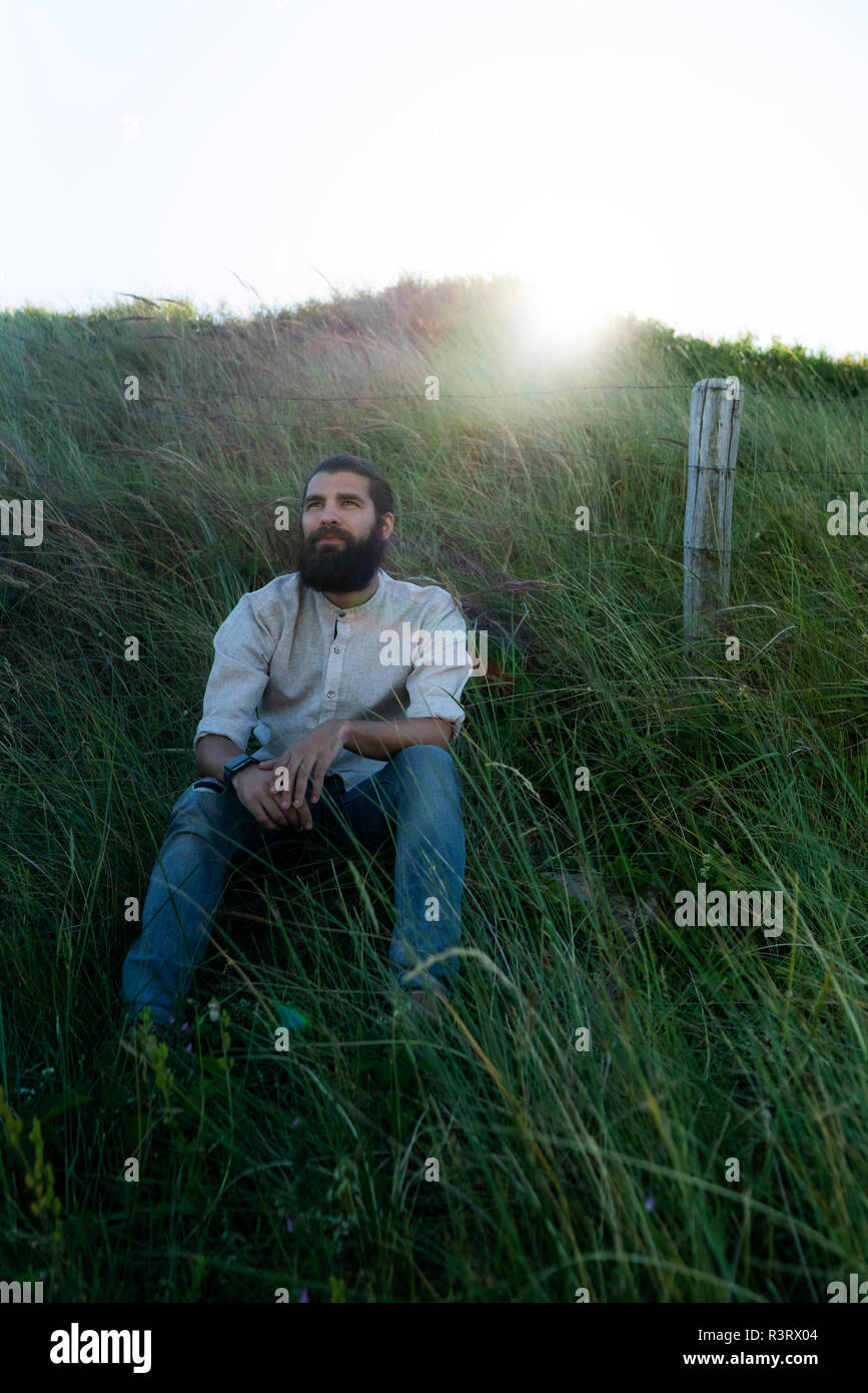 Giovane uomo seduto sull'erba nelle dune, fantasticando Foto Stock