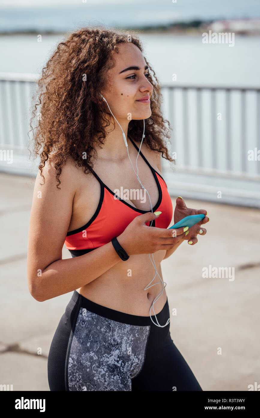 Sorridente atletica giovane donna con lo smartphone e gli auricolari al Riverside Foto Stock