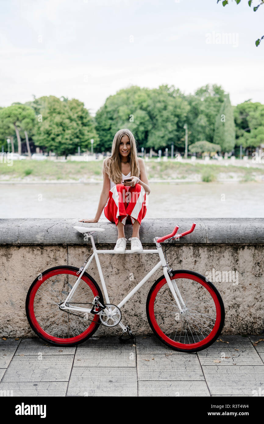 Ragazza con un telefono cellulare seduti in riva al fiume e prossimo alla bicicletta Foto Stock