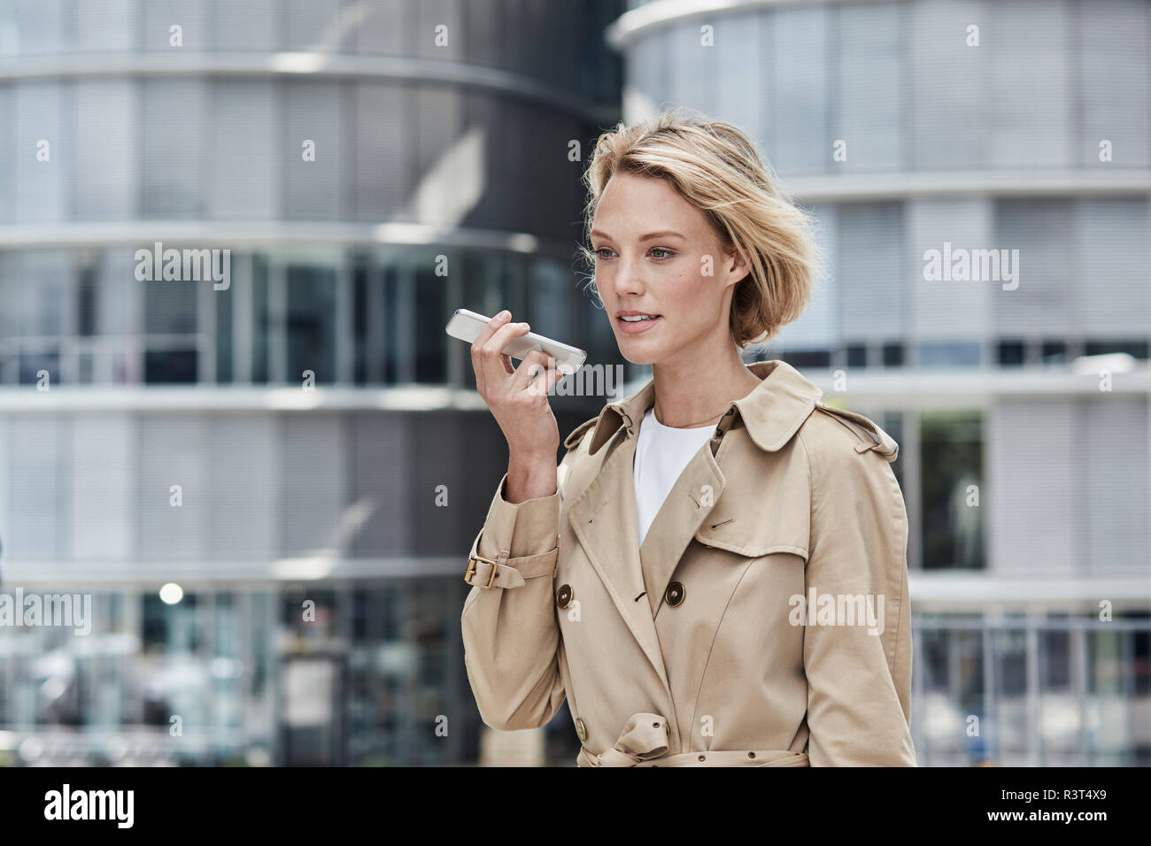 Germania, Duesseldorf, ritratto del biondo imprenditrice indossando il beige trench utilizza lo smartphone Foto Stock