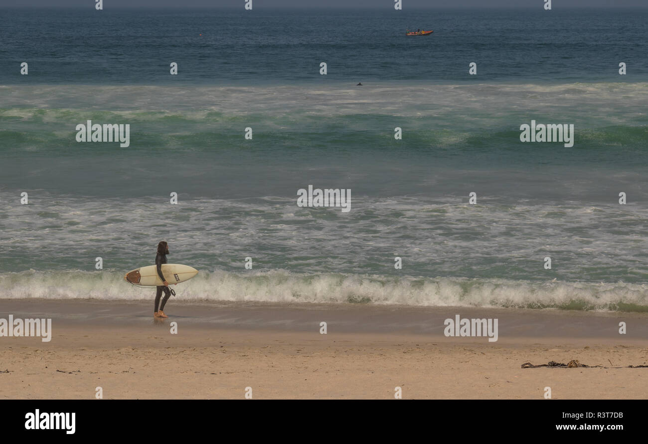 Cape Town, Sud Africa - un surfista non identificato passeggiate lungo la spiaggia di Bloubergstrand su un ventoso e nebbioso giorno immagine con spazio di copia Foto Stock