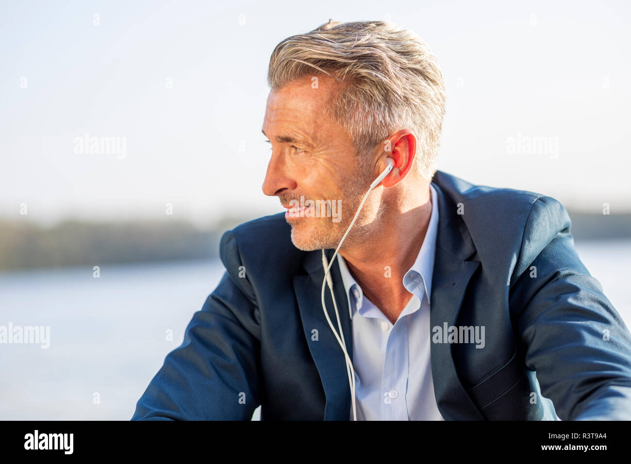 Imprenditore sorridente ascoltando la musica con gli auricolari al lago Foto Stock