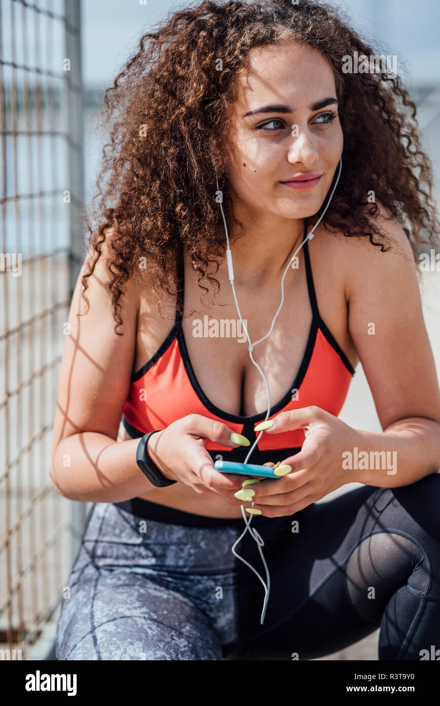 Sorridente atletica giovane donna con lo smartphone e auricolari accovacciato all'aperto Foto Stock