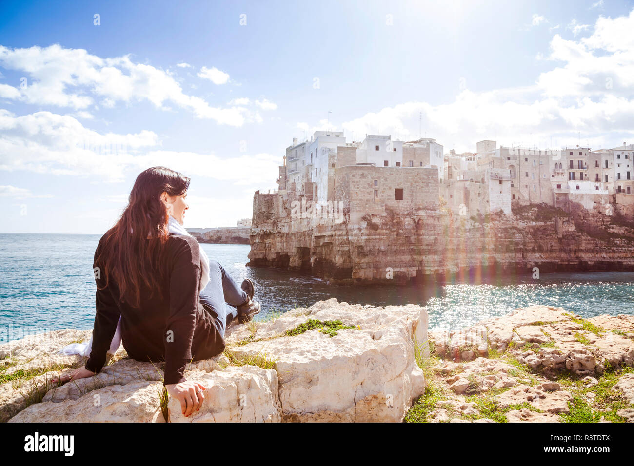 L'Italia, Puglia, Polognano a Mare, vista posteriore della donna rilassante sulle rocce guardando il centro storico Foto Stock