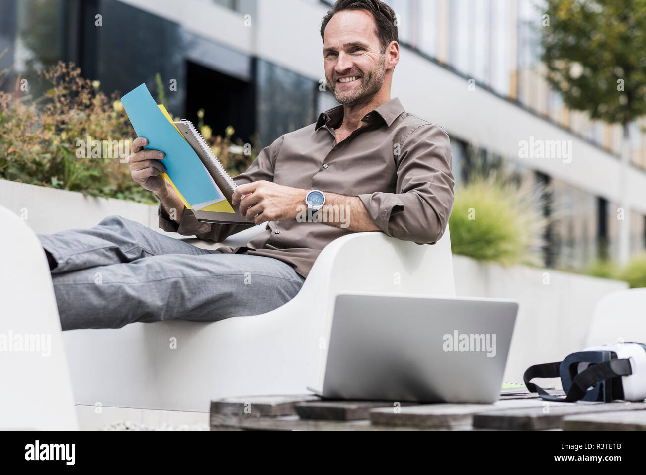 Imprenditore sorridente con il documento, laptop e occhiali per realtà virtuale seduti sulla terrazza Foto Stock