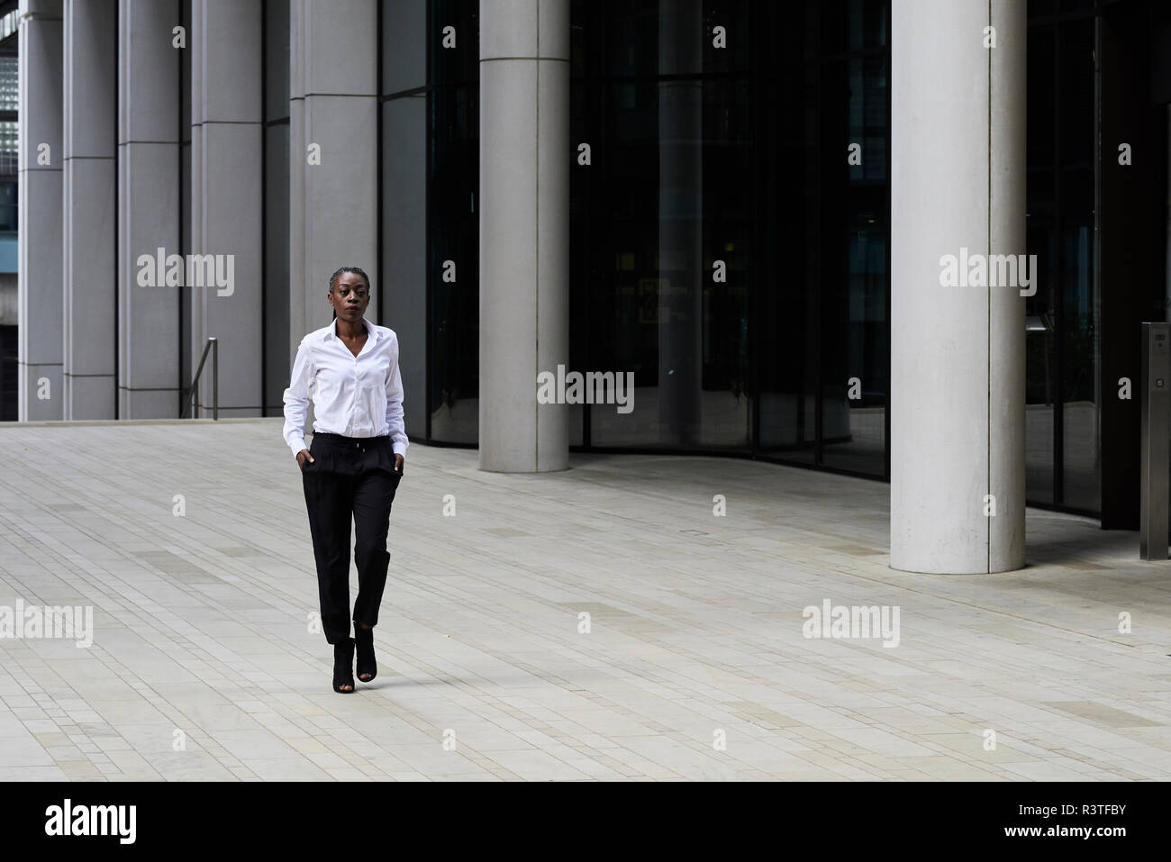 Imprenditrice che indossa una camicia bianca e pantaloni neri a piedi nella parte anteriore di un moderno edificio per uffici Foto Stock