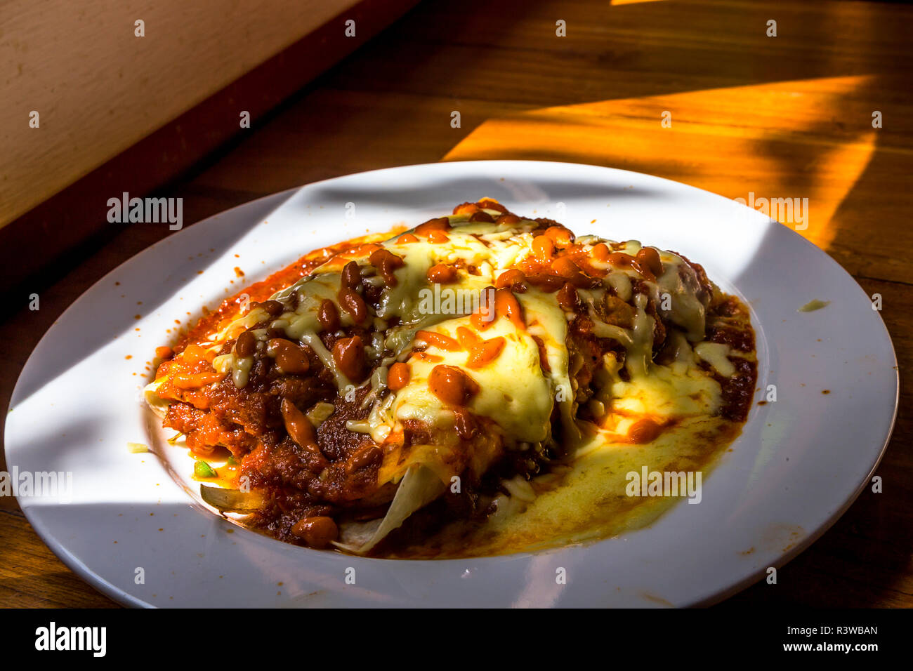 Enchilada servita con formaggio e salsa di peperoncino il riempimento di una piastra bianca con elevate aree illuminate e ombre su una tavola di legno. Foto Stock