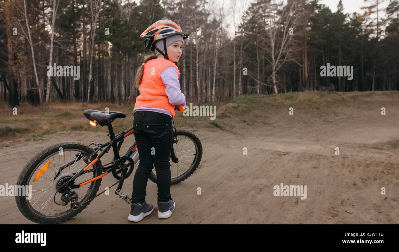 Uno caucasico giostre per bambini bike via strada nel parco di sporco. Ragazza a cavallo nero ciclo arancione in pista. Kid va di fare sport bicicletta. Biker moto ride Foto Stock