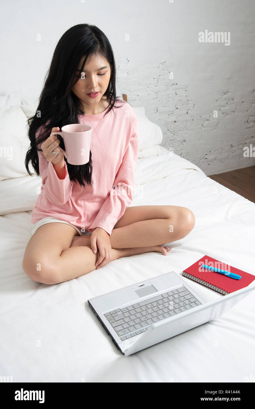 Donna seduta sul letto ha tazza di caffè in mano, rilassante guardando un computer notebook. Foto Stock