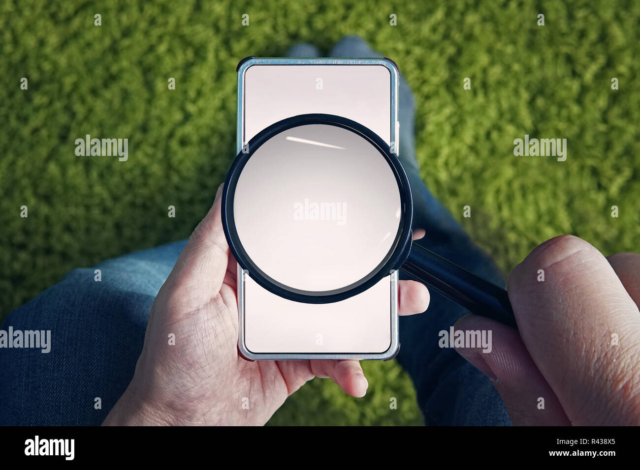 Vista dall'alto di uomo seduto con lo smartphone in bianco e lente di ingrandimento sopra il suo schermo. L'uomo detiene sulla lente di ingrandimento per trovare qualcosa di internet mobile. Uso del telefono cellulare per la ricerca di informazioni in linea. Foto Stock